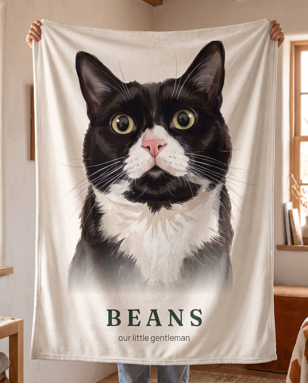A warm white throw with Beans’s tuxedo cat portrait and name, held up to show the full design.